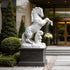 White marble rearing horse statue displayed at a luxury outdoor entrance with shrubs, stone steps, and modern building background.
