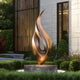 Abstract large dancing flame bronze sculpture in a garden setting with a building in the background