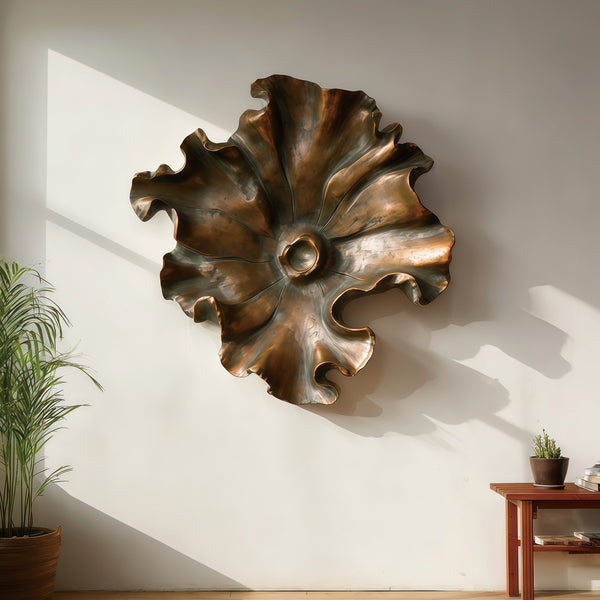 Bronze wall sculpture sunlit ripples floral form on a white wall with a plant and wooden shelf in the background