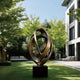 Continuous Interlocking Ribbon Abstract Bronze Sculpture on a black pedestal base, set in a modern outdoor courtyard with green grass, trees, and contemporary white buildings.
