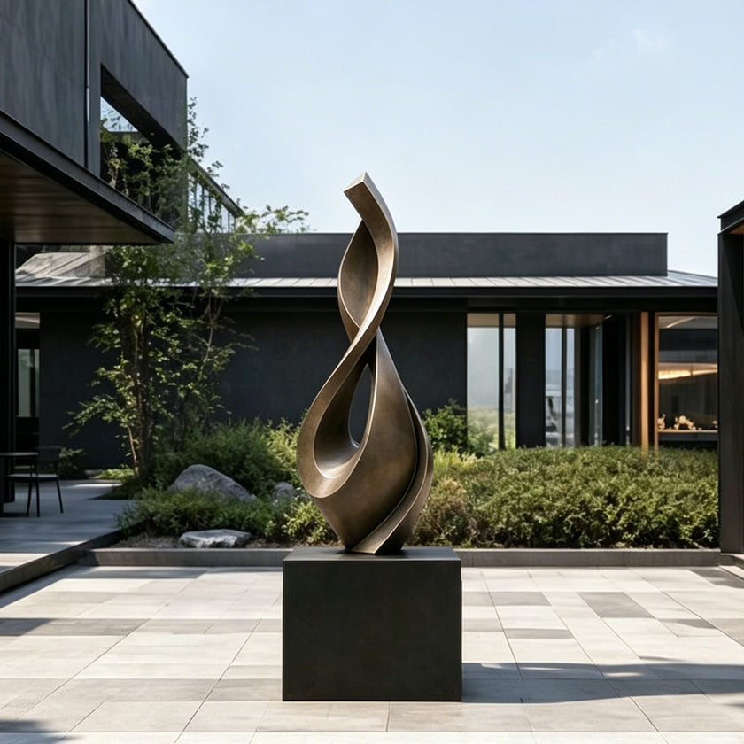 Fluid Twisting Infinity Teardrop Bronze Landscape Sculptures on a dark rectangular pedestal in an outdoor courtyard setting with modern dark buildings, plants, and a tiled patio.