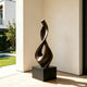 Fluid Twisting Infinity Teardrop Bronze Landscape Sculptures with a flowing, intertwined looped form mounted on a black rectangular pedestal, displayed in a modern interior space with light wood flooring and a sunlit cream wall.