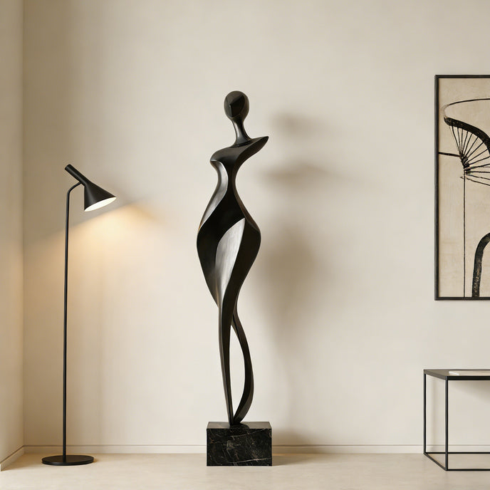 Rhythmic Walking Woman Abstract Bronze Figure Sculpture of a stylized female figure standing on a rectangular black marble base, displayed in a minimalist room next to a lit floor lamp and a framed abstract print.