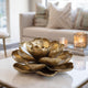 Blooming Golden Lotus Tabletop Floral Bronze Sculpture sits on a light marble coffee table in a modern living room setting, with a beige sofa and decorative candles blurred in the background.