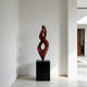 Twisting Red Bronze Flame Floor Sculpture Abstract Art Sculptures of two intertwined spiral forms in a glossy dark reddish-brown finish, mounted on a black rectangular pedestal, set against a minimalist white interior wall.