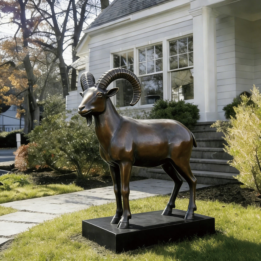 Classic Mountain Ram Outdoor Sculpture Bronze Statuary with large curved horns stands on a black rectangular base in a landscaped yard in front of a white house.