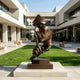 A Silent Watcher Head Sculpture Bronze Outdoor Statuary of a stylized human head resting on a rectangular pedestal, placed in an outdoor modern plaza with green grass, walkways, and contemporary buildings in the background.