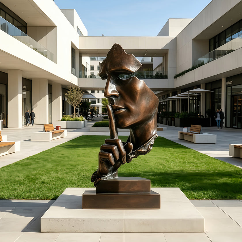 A Silent Watcher Head Sculpture Bronze Outdoor Statuary of a stylized human head resting on a rectangular pedestal, placed in an outdoor modern plaza with green grass, walkways, and contemporary buildings in the background.
