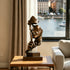 Silent Watcher Head Sculpture Bronze Outdoor Statuary of a stylized face holding a phone to its lips, displayed on a wooden side table in a modern living room setting with a window overlooking a waterfront.