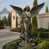 Classic Winged Guardian Angel Bronze Sculpture Garden Large Statues with outstretched wings stands on a circular base in a landscaped garden courtyard with trimmed hedges, a paved walkway, and a stone gate in the background.