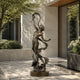 Elegant Dancing Woman Bronze Statue Outdoor Female Figure Sculpture of a nude female figure with flowing drapery stands on a circular base in an outdoor courtyard setting with stone walls, trees, and plants.