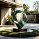 Twisted Emerald Ribbon Abstract Art Sculptures Modern Contemporary Sculpture of two intertwined curved forms in a deep green metallic finish, mounted on a black rectangular pedestal. The sculpture is situated outdoors on a circular landscaped planter bed with manicured greenery, set against a contemporary building facade with trees in the background.