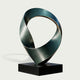 Twisted Emerald Ribbon Abstract Art Sculptures Modern Contemporary Sculpture with a twisted ribbon-like form in dark metallic teal and gray, mounted on a black rectangular base against a light gray background.