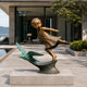 Dancing Girl And Flying Bird Bronze Figure Sculpture Outdoor Sculpture. A child in a dress flying above a green patinated bird mounted on a wave-like base, displayed on a stone pedestal in a modern outdoor patio setting.