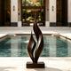 Dancing Twin Flames Bronze Art Sculpture with two tall, flowing leaf-like forms on a rectangular base, placed in front of a swimming pool in a luxurious outdoor courtyard.