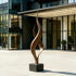 Dual Weave Abstract Bronze Sculpture with two flowing spiral forms rising from a dark rectangular base, displayed outdoors in front of a contemporary glass-and-steel building on a light paved plaza.