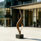 Dual Weave Abstract Bronze Sculpture with two flowing spiral forms rising from a dark rectangular base, displayed outdoors in front of a contemporary glass-and-steel building on a light paved plaza.
