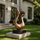 Large abstract bronze sculpture with flowing organic curves and openwork forms, displayed on a black pedestal in the landscaped garden of a modern luxury villa.
