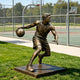 Life-size bronze basketball player sculpture in a dynamic dribbling pose, featuring detailed athletic musculature, basketball uniform, sneakers, and ball, displayed on a square base beside an outdoor basketball court.