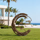 Abstract bronze crescent garden sculpture installed on a green lawn at a coastal villa, with textured sweeping branches and delicate floating leaves framed by palm trees, modern architecture, and an ocean view.