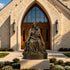 Crowned bronze Madonna and Child sculpture displayed outside a church entrance, depicting the seated Virgin Mary tenderly holding the infant Jesus, with flowing robes and an ornate floral pedestal base.