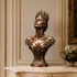 Crowned bronze queen bust sculpture displayed on a marble console in a luxurious classical interior, featuring an upward-facing female portrait, elaborate Gothic-style crown, graceful shoulders, ornate botanical scrollwork, and warm bronze patina.