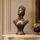 Crowned bronze queen bust sculpture displayed on a marble console in a luxurious classical interior, featuring an upward-facing female portrait, elaborate Gothic-style crown, graceful shoulders, ornate botanical scrollwork, and warm bronze patina.