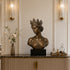 Crowned bronze queen bust sculpture displayed on a dark marble console in an elegant luxury interior, featuring a serene downward gaze, detailed floral crown, swept-back hair, off-shoulder drapery, and rich antique bronze patina.