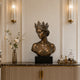 Crowned bronze queen bust sculpture displayed on a dark marble console in an elegant luxury interior, featuring a serene downward gaze, detailed floral crown, swept-back hair, off-shoulder drapery, and rich antique bronze patina.