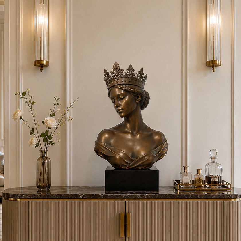 Crowned bronze queen bust sculpture displayed on a dark marble console in an elegant luxury interior, featuring a serene downward gaze, detailed floral crown, swept-back hair, off-shoulder drapery, and rich antique bronze patina.