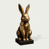 Faceted bronze rabbit sculpture in a seated position with long upright ears, polygonal geometric body, angular facial details, warm bronze highlights, antique patina, and a square dark pedestal base.