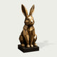 Faceted bronze rabbit sculpture in a seated position with long upright ears, polygonal geometric body, angular facial details, warm bronze highlights, antique patina, and a square dark pedestal base.