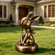 Charming bronze rabbit sculpture displayed on a green lawn outside an elegant residence, featuring closed eyes, long upright ears, a butterfly resting on its nose, a tied neck scarf, seated pose, rounded pedestal, and warm antique bronze patina.