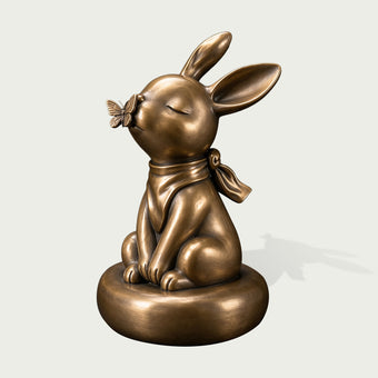 Antique bronze rabbit sculpture seated on a rounded base with eyes gently closed, long expressive ears, a detailed butterfly touching its nose, tied scarf around the neck, smooth rounded forms, and softly highlighted bronze surface.