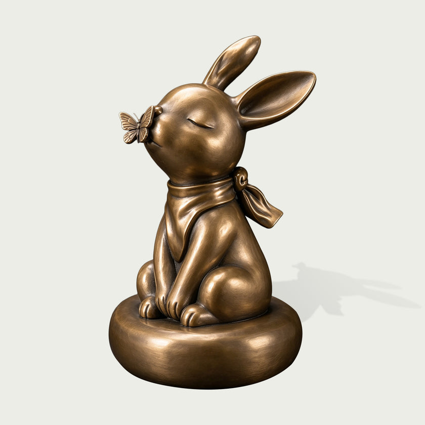 Antique bronze rabbit sculpture seated on a rounded base with eyes gently closed, long expressive ears, a detailed butterfly touching its nose, tied scarf around the neck, smooth rounded forms, and softly highlighted bronze surface.