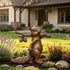 Whimsical bronze squirrel garden sculpture carrying a textured tree branch over its shoulder with two hanging acorns, displayed among lavender and flowering plants in a landscaped residential garden, with a warm antique bronze patina and detailed bushy tail.