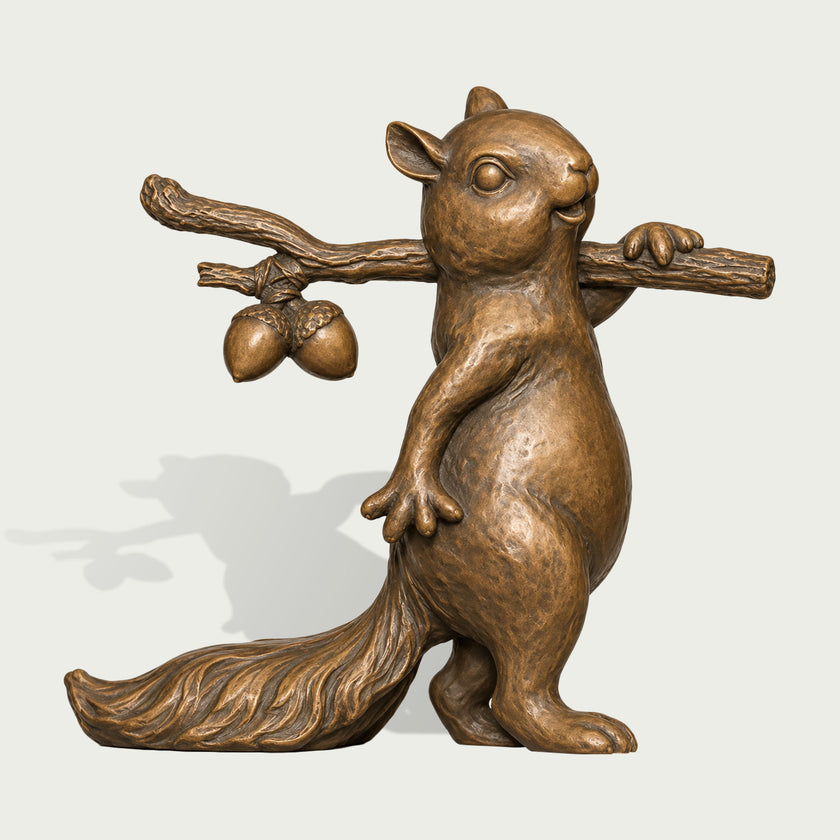 Full product view of a bronze squirrel sculpture carrying a textured tree branch across its shoulder with two sculpted acorns, featuring a playful upright pose, rounded belly, expressive facial details, large bushy tail, and hand-textured antique bronze surface.