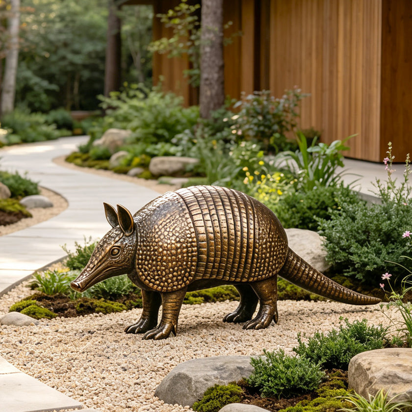 Bronze armadillo sculpture displayed in a landscaped garden beside a curved walkway, featuring a long tapered tail, pointed ears, elongated snout, textured armor plates, detailed claws, and a warm antique bronze patina.