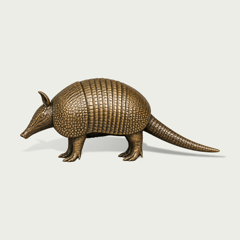 Side view of an antique bronze armadillo animal sculpture with a rounded armored body, individually textured shell plates, pointed snout and ears, sturdy clawed legs, and a long segmented tail.