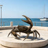 Large bronze crab sculpture displayed on a circular stone platform by the sea, featuring oversized raised claws, articulated legs, a textured shell, detailed joints, and a rich antique bronze surface against a coastal marina setting.