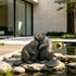 Bronze sculpture of two embracing beavers displayed on a natural stone beside a landscaped garden pond, featuring detailed fur, broad textured tails, gentle expressions, and weathered green patina accents in a modern outdoor setting.