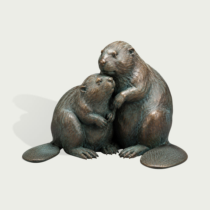 Bronze beaver pair sculpture depicting two affectionate beavers sitting closely together, with one gently embracing the other, detailed textured fur, broad patterned tails, expressive faces, and natural green patina accents over warm bronze.