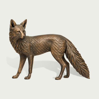 Freestanding bronze fox animal sculpture shown against a neutral background, with a graceful standing pose, turned head, pointed ears, detailed layered fur, slender legs, and a large flowing bushy tail.