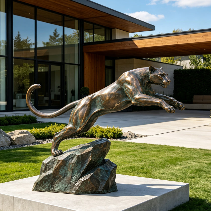 Large bronze leaping panther sculpture displayed in a modern residential garden, featuring an athletic body in midair, extended front legs, a long curved tail, and a rugged rock-form base with aged green patina accents.