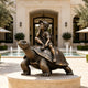 Whimsical bronze sculpture of a smiling boy wearing a cap and backpack while sitting on a giant tortoise, displayed on a circular stone pedestal beside a luxury courtyard fountain.