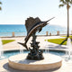 Monumental bronze sailfish sculpture displayed on a circular stone pedestal beside an outdoor fountain overlooking the ocean, with a dynamic jumping pose, raised dorsal fin, textured wave base, and dark antique bronze patina.
