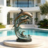 Large bronze dolphin sculpture displayed beside a luxury swimming pool, featuring two dolphins leaping above curling ocean waves with smooth sculpted bodies, warm bronze highlights, and blue-green verdigris patina.