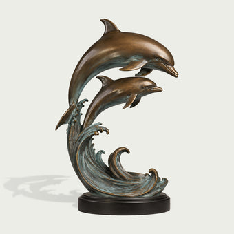 Bronze sculpture of two leaping dolphins rising above stylized curling ocean waves, detailed with streamlined bodies, sculpted fins, warm antique bronze tones, blue-green verdigris patina, and a round dark base.