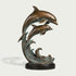 Bronze sculpture of two leaping dolphins rising above stylized curling ocean waves, detailed with streamlined bodies, sculpted fins, warm antique bronze tones, blue-green verdigris patina, and a round dark base.
