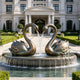 Bronze swan pair fountain sculpture displayed in a formal villa garden, featuring two elegant swans with curved necks forming a heart shape, detailed layered feathers, green patina accents, and water flowing from their beaks.